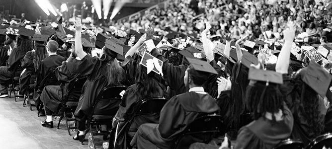 Black & white photo of students and crowd at graduation ceremony; two women in center are cheering.