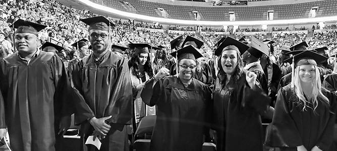 Black & white photo of students and crowd at graduation ceremony; two women in center are cheering.